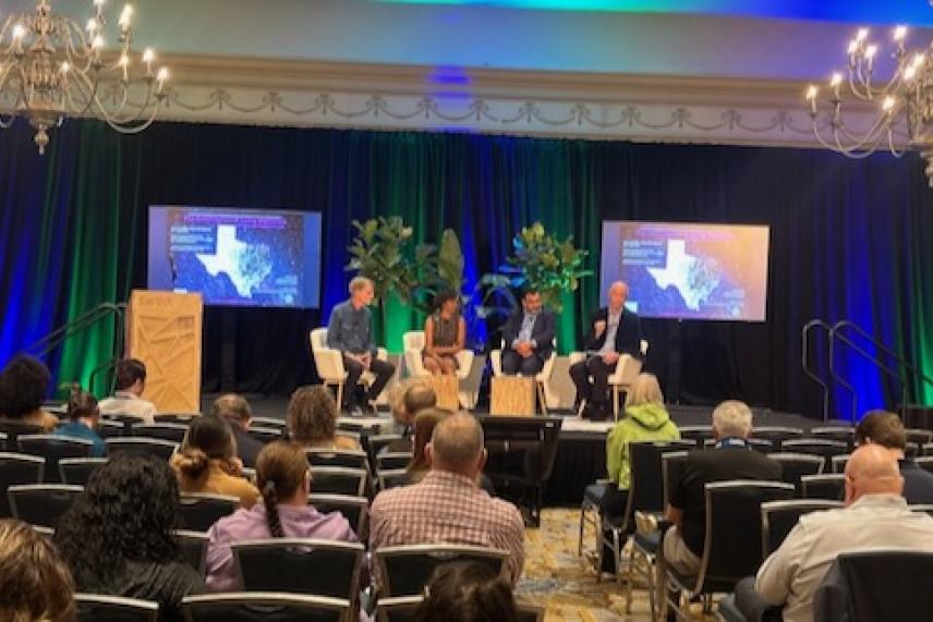 Panel discussion in a conference room with two screens and chandeliers overhead.
