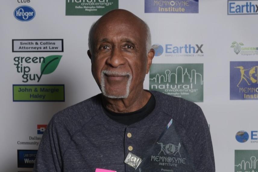 Elderly man smiling at a formal event, standing against a backdrop with logos.