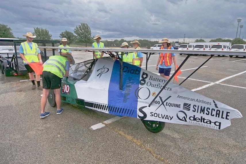 Small car covered in logos with people in safety vests around it.