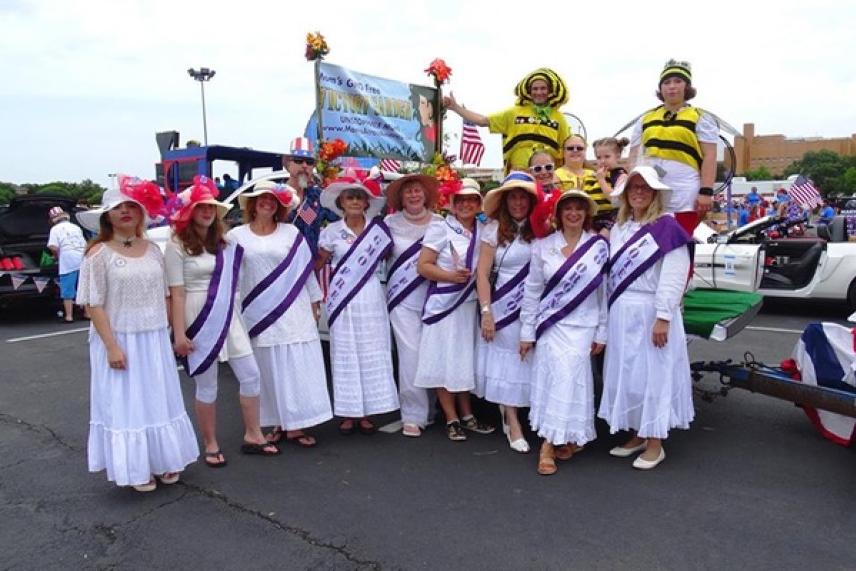 Women in white dresses and hats pose in front of a float at an outdoor event.