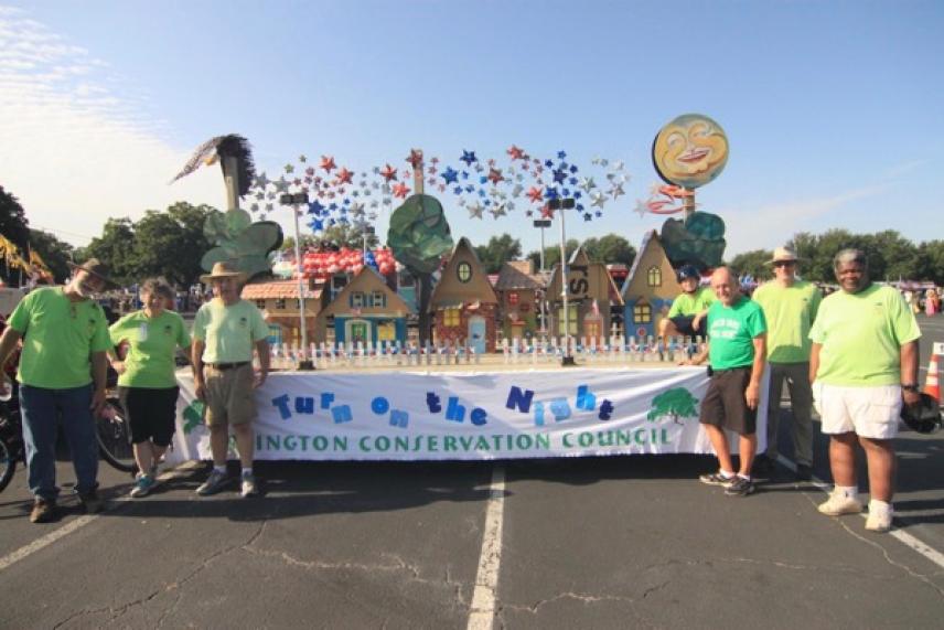 People in green shirts stand beside a colorful float in a parking lot.