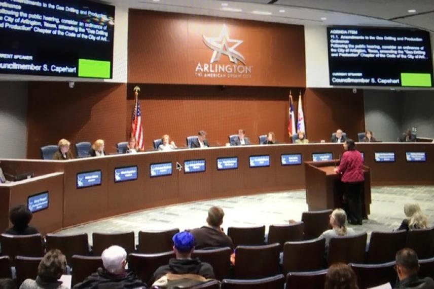 Council meeting in session, a person speaks at a podium, attendees seated.