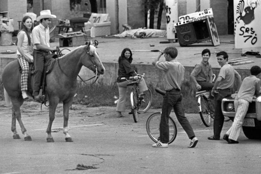 Crowded street with people on horseback, bikes, and standing around. Black and white photo.