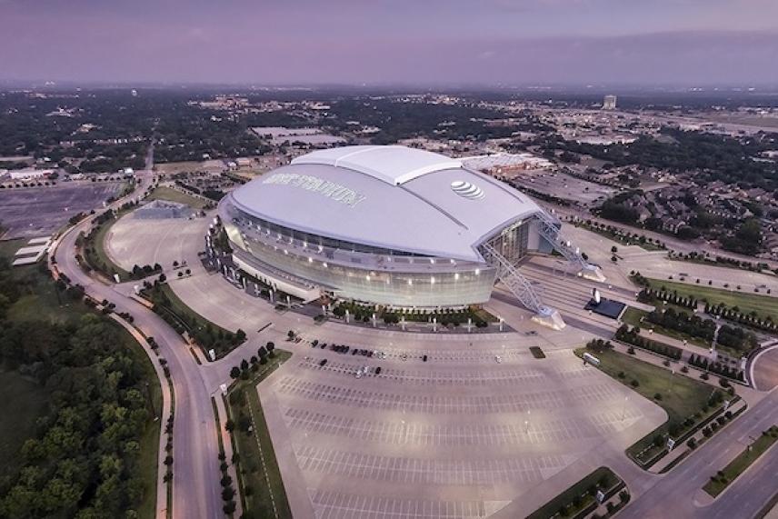 Large stadium with a domed roof at dusk, surrounded by parking lots and cityscape.