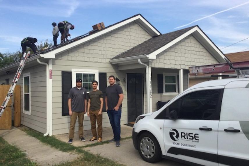 Workers installing solar panels on a house roof; a van with a logo is parked nearby.