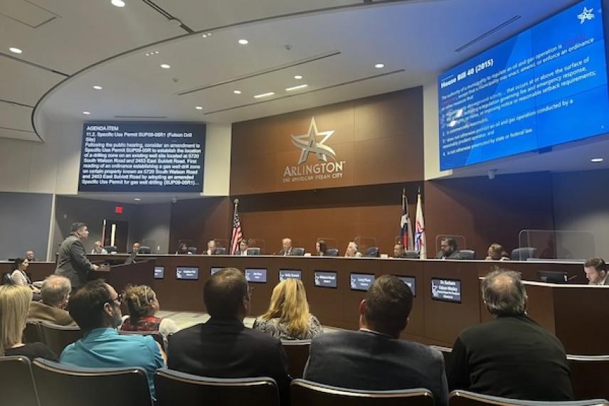 Council meeting in session, people seated, presentation on screens.