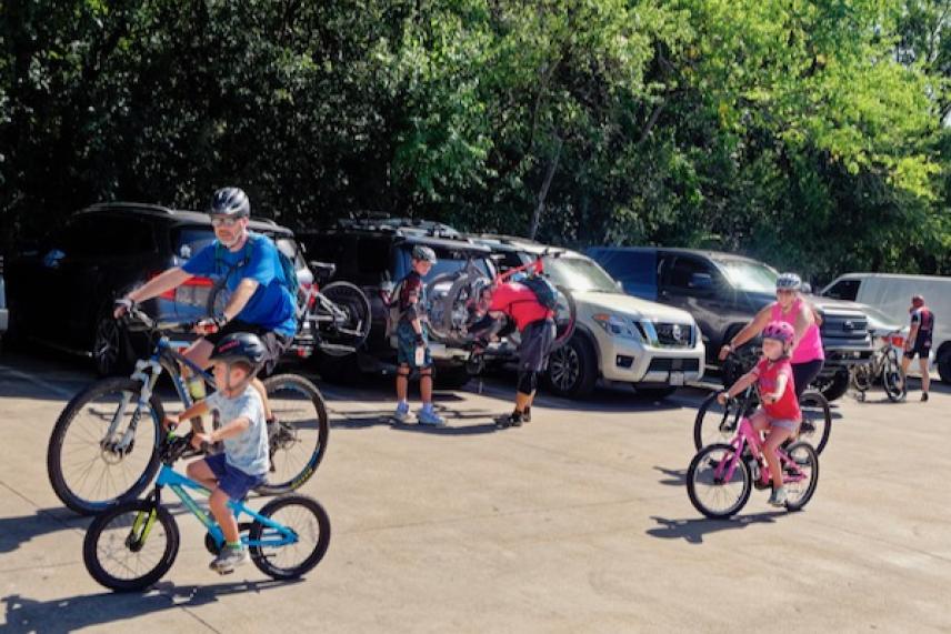 Families preparing bicycles in a sunny parking lot near greenery.