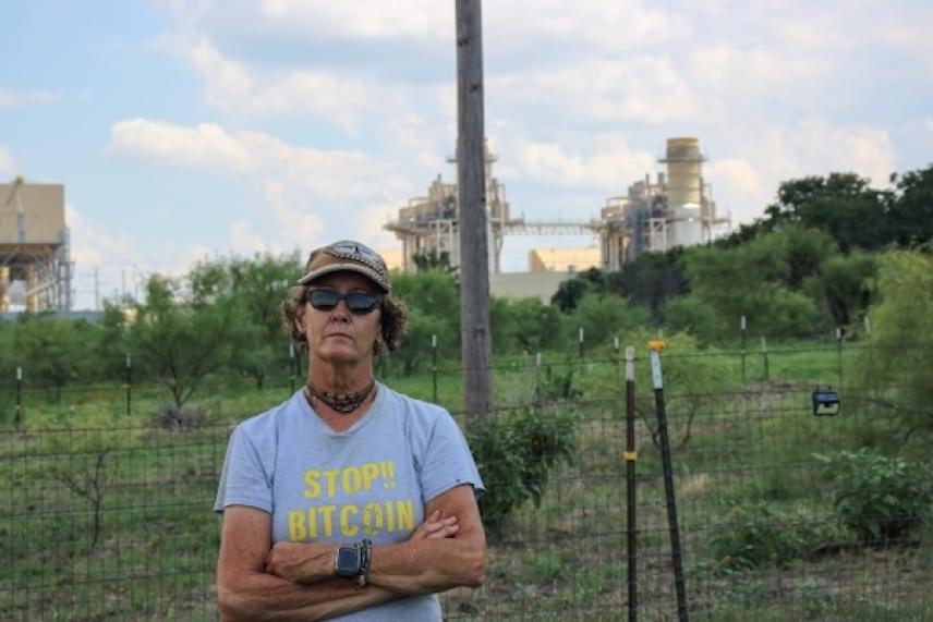 Person in a hat stands with arms crossed in a field with industrial structures behind.