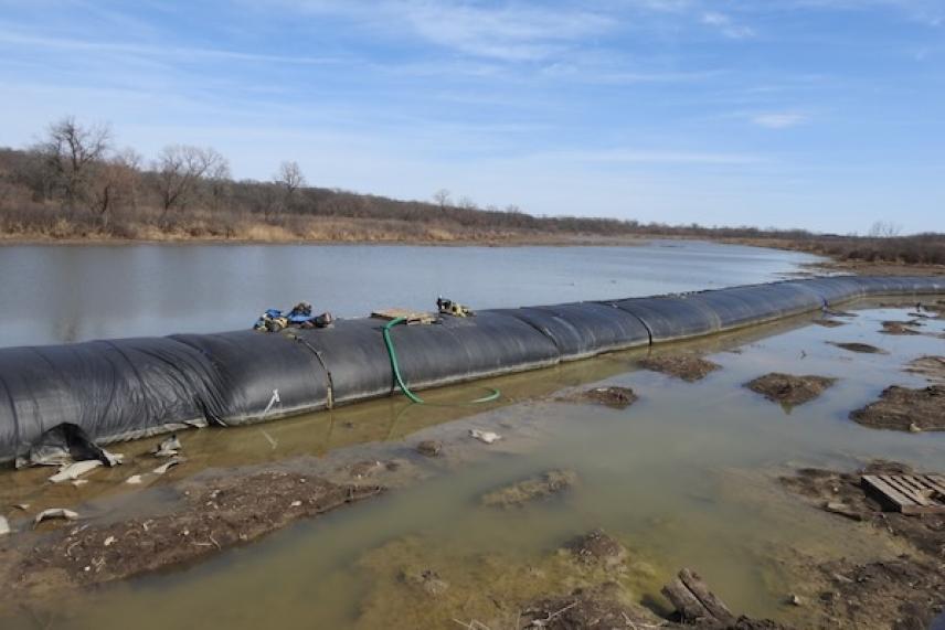 Large black inflatable barrier along a riverbank on a clear day.