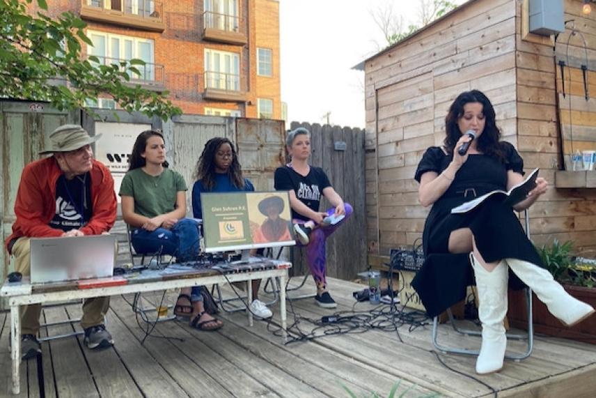Outdoor panel discussion with five people seated, one reading from a book.