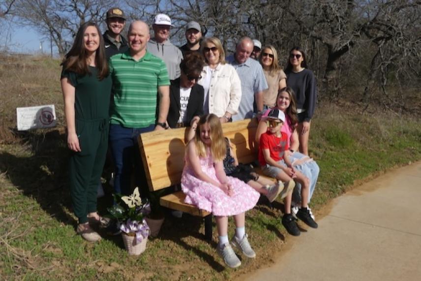 Family poses outdoors around a bench, children sitting, some carrying flowers, clear sky.