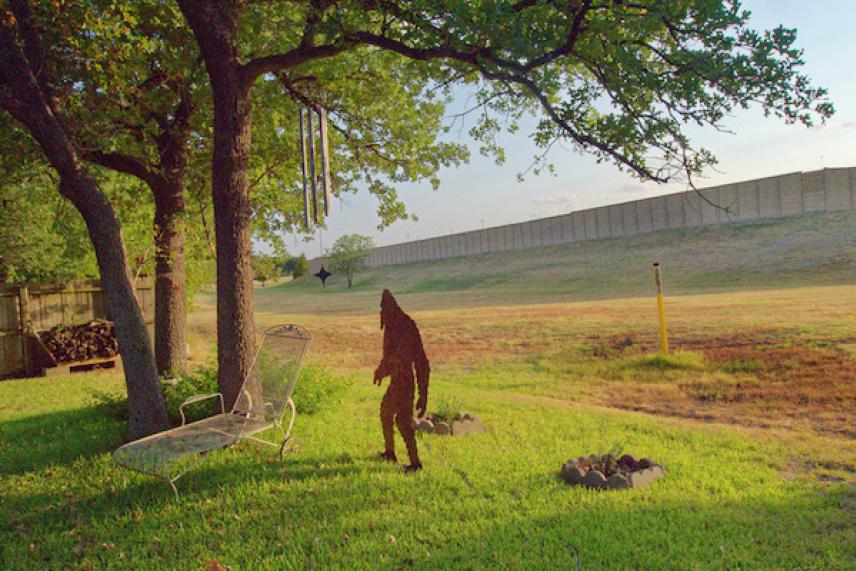 A person in a brown costume stands under a tree with a wind chime on a grassy field.