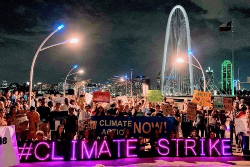 Crowd at night with neon "Climate Strike" sign, cityscape backdrop.