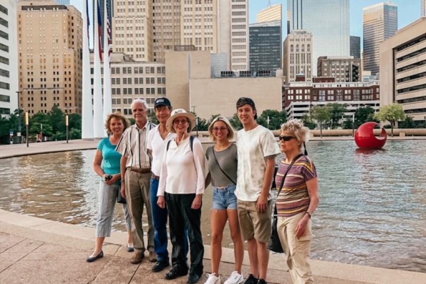 Group of seven people smiling in front of a city fountain and skyscrapers.