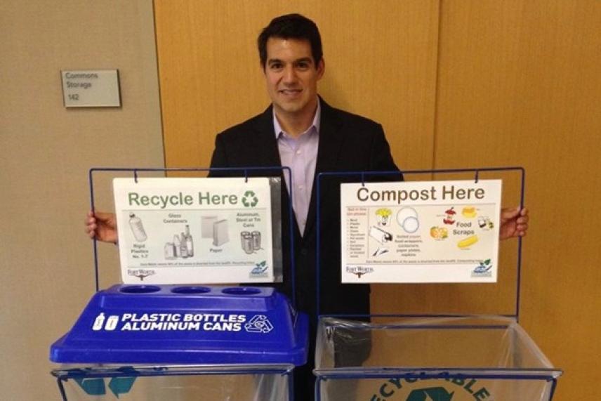 Man holding recycle and compost signs above bins indoors.