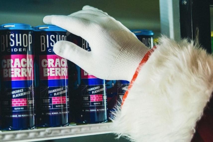 Santa's gloved hand reaching for a Bishop cider can on a shelf.