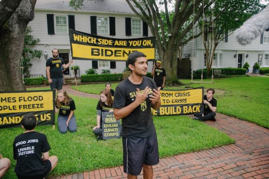 Protesters in black shirts with signs on a lawn near a white house.