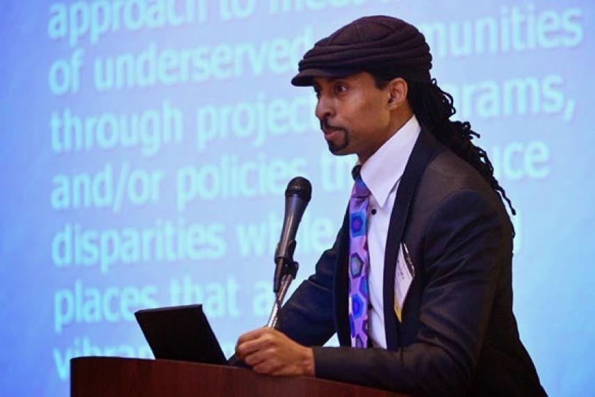 Man in suit and cap speaking at a podium with projected text in the background.
