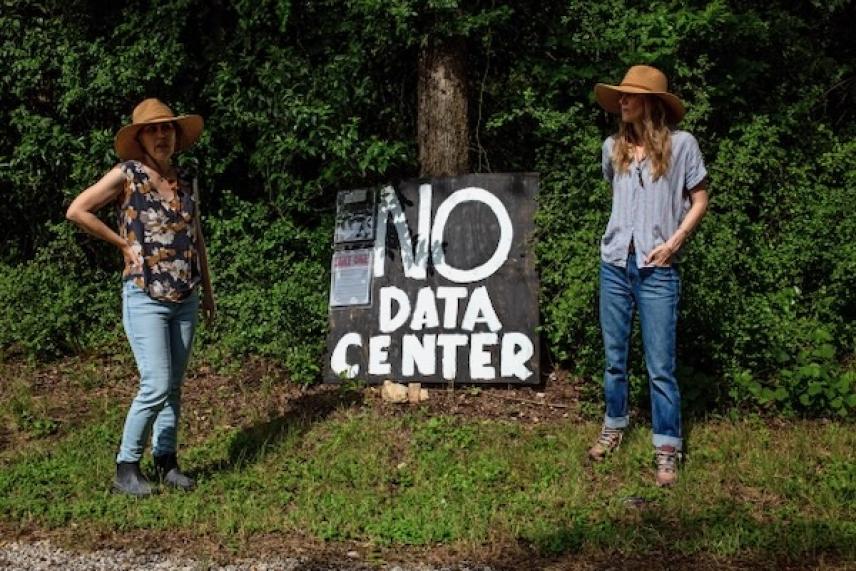 Two people in hats stand beside a "No Data Center" sign near trees.