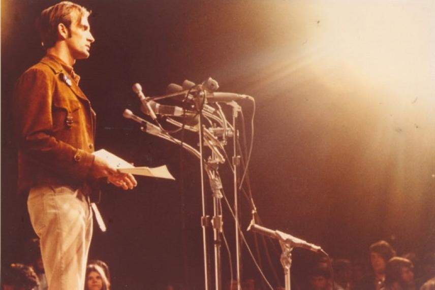 Man speaking at podium with multiple microphones, warm lighting, audience visible.
