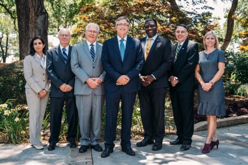 Seven people in formal attire standing outdoors, smiling.