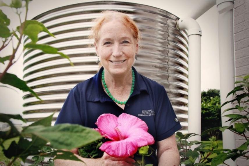 Woman smiling, holding a large pink flower, with a water tank in the background.