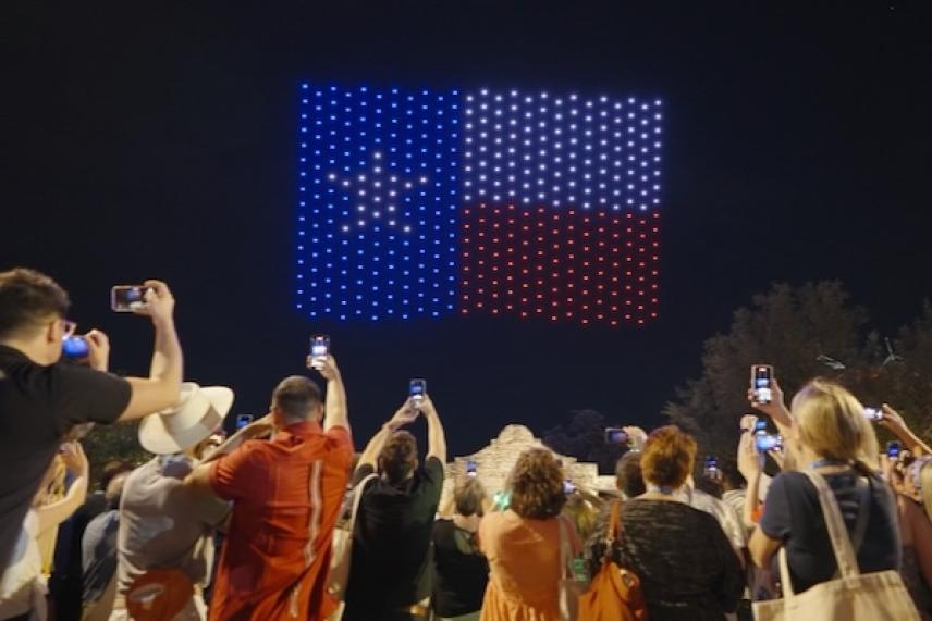 Crowd watches Texas flag light display at night, taking photos.