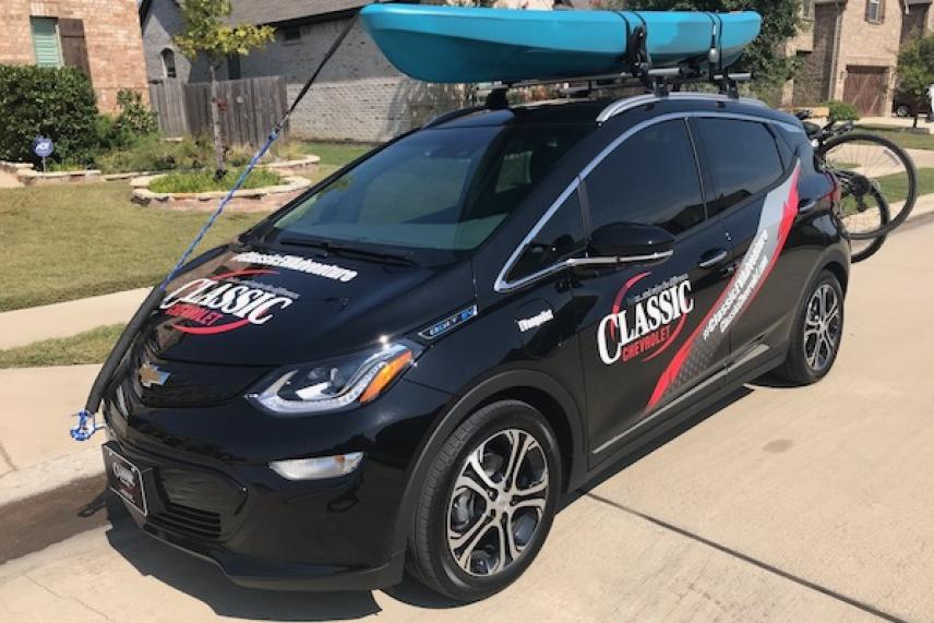 Black car with a blue kayak on roof racks, parked in a suburban driveway.