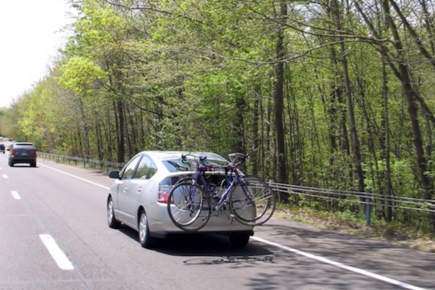 Silver car with bicycles on back driving on tree-lined road.