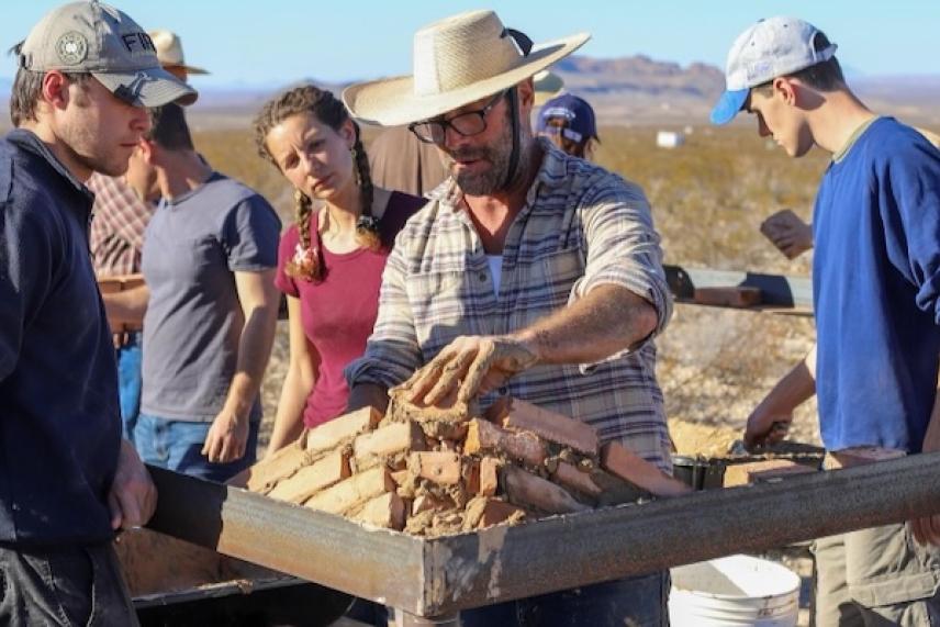 Group examines artifacts in desert setting. Man in straw hat leads activity.