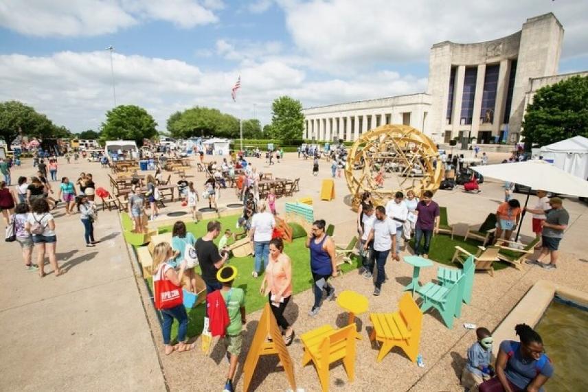 Crowded outdoor festival with colorful seating and people mingling under a blue sky.