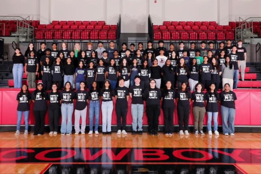 Large group of students wearing black shirts, standing in gymnasium bleachers.