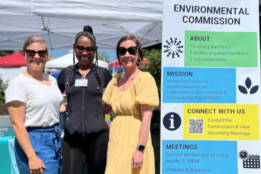 Three women smiling beside an Environmental Commission sign at an outdoor event.