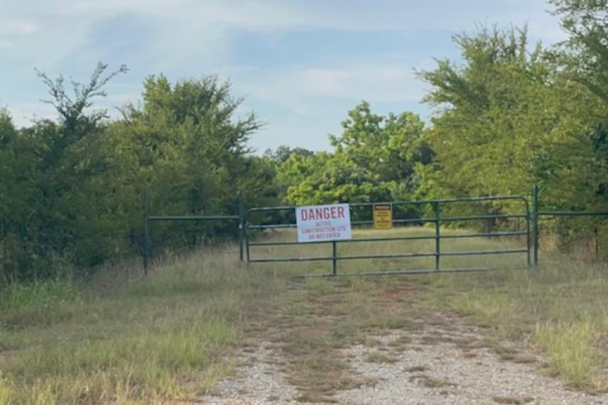 Gravel path leading to a gated entrance with warning signs, surrounded by trees.