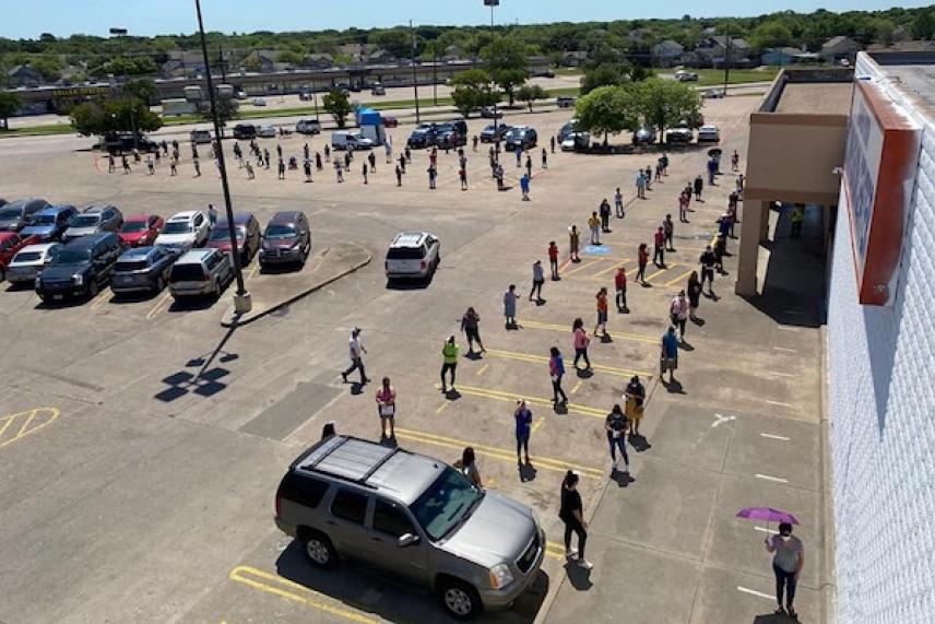People lined up outside a building, maintaining distance, on a sunny day.