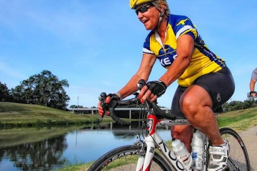 Cyclist in yellow jersey rides by a river, smiling under a clear blue sky.