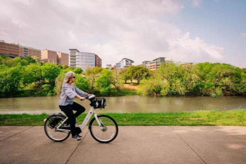 Person biking on a riverside path with cityscape and greenery in the background.