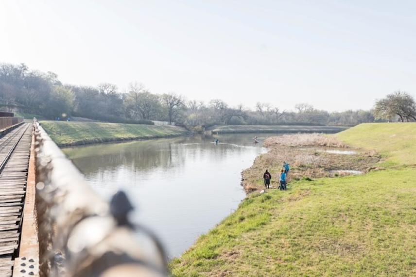 Riverbank with grassy hills, two people near the water, railway bridge on the left.