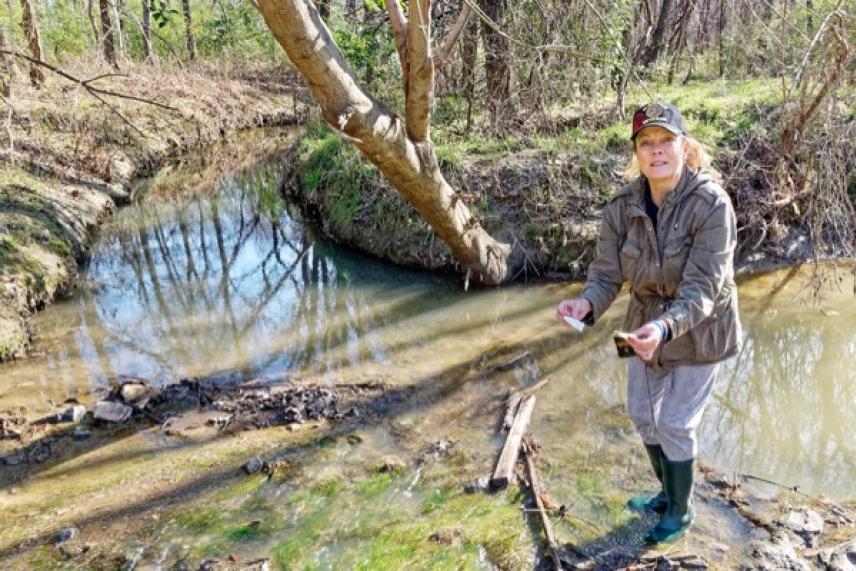 Woman in a cap stands by a creek, holding a small object, surrounded by trees.