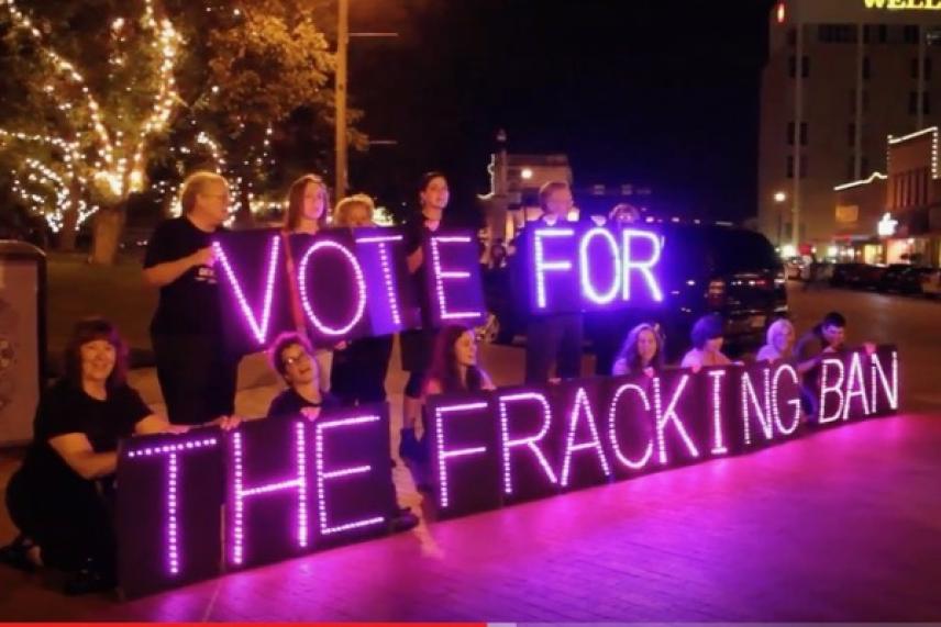 Group holding illuminated signs reading "Vote for the Fracking Ban" at night.