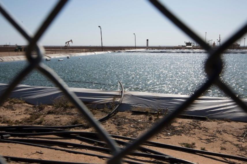 Chain-link fence view of a sparkling water reservoir and distant oil pumps.