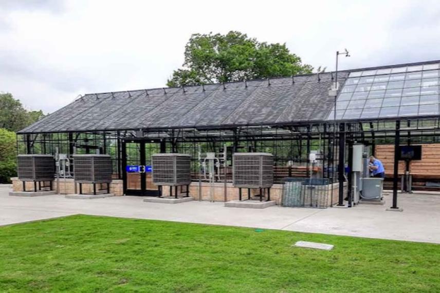 Glass greenhouse with large air conditioning units outside, set on a concrete path and green grass.