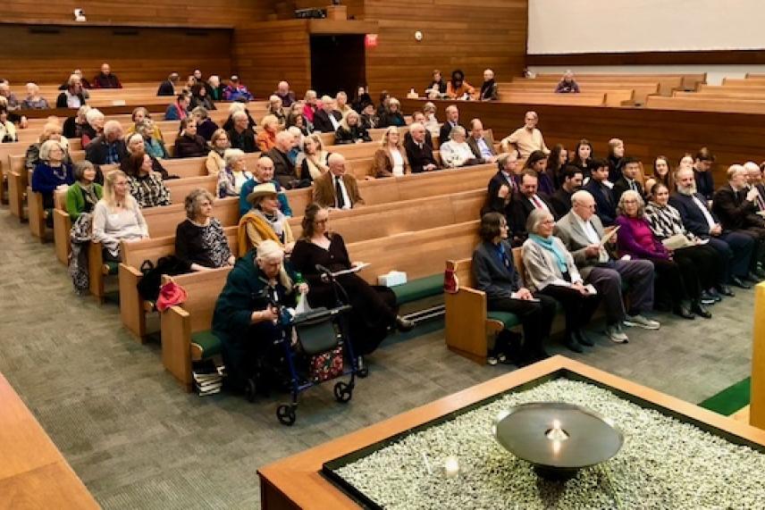 Audience seated in a wooden church with an altar in the foreground.