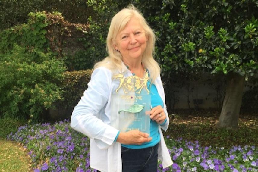 Blonde woman smiling, holding a clear trophy, in a garden with flowers and greenery.