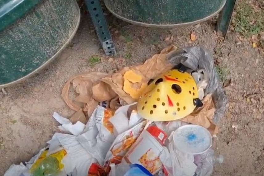 Trash with a yellow hockey mask near green bins on dirt ground.