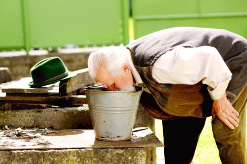 Elderly man drinking from a metal bucket outdoors.