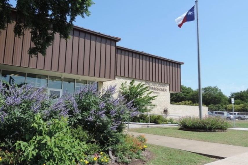 Modern building with Texas flag, surrounded by greenery and a clear blue sky.