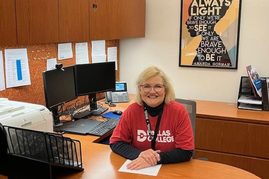 Smiling woman in a red shirt sits at a desk in an office with dual monitors.