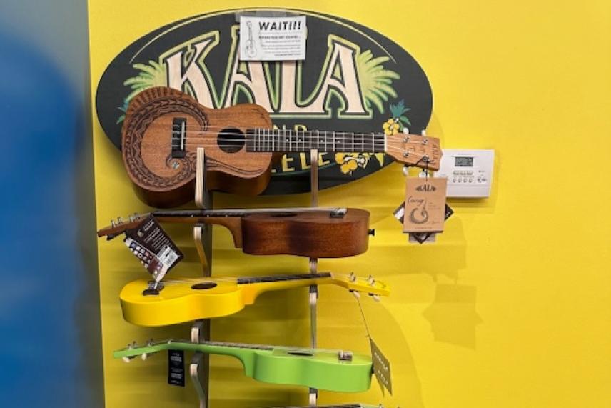 Ukuleles in various colors displayed on a yellow wall with a logo above.