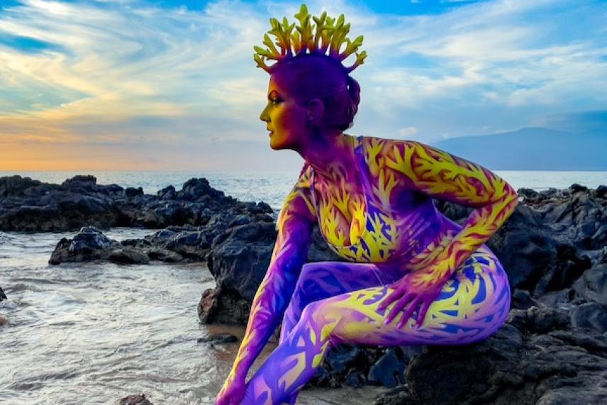 Colorfully painted person with crown, sitting on rocks by the sea under a vibrant sky.